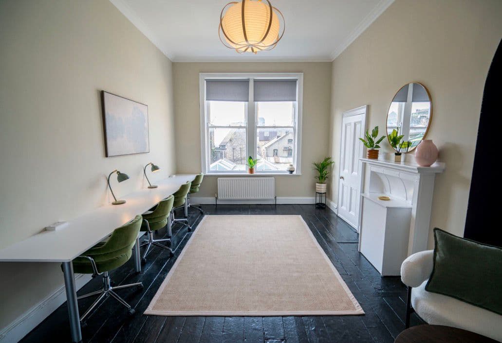 A bright workspace with a run of desks, green chairs and a restored fireplace